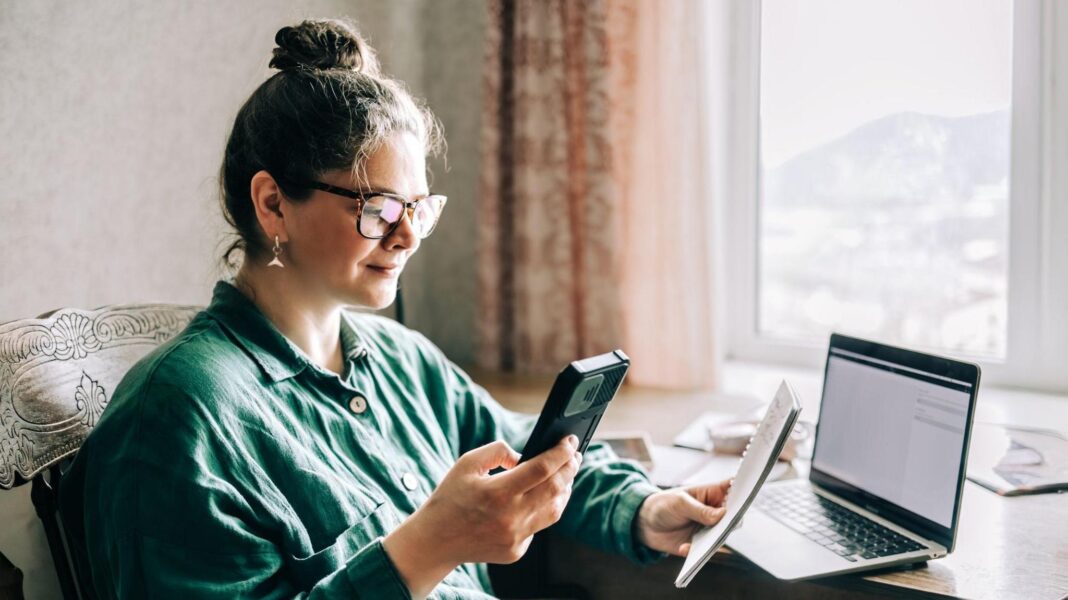 The £70 refund letter that isn’t a scam Woman sits at a window seat holding a phone and a pad with a computer on a table in front of her.