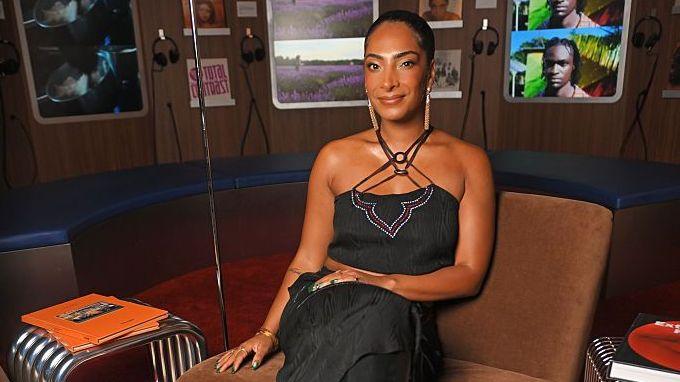 Priya wears a strappy black dress with a purple design and long gold patterned earrings. She has black hair, which is scraped back in a tight ponytail style.