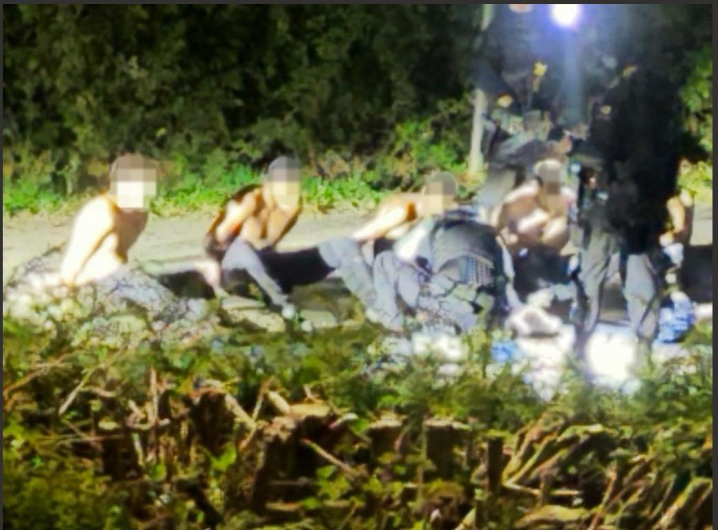 Detained men in Whelford. Their faces are blurred. They are shirtless. Trees line the background.