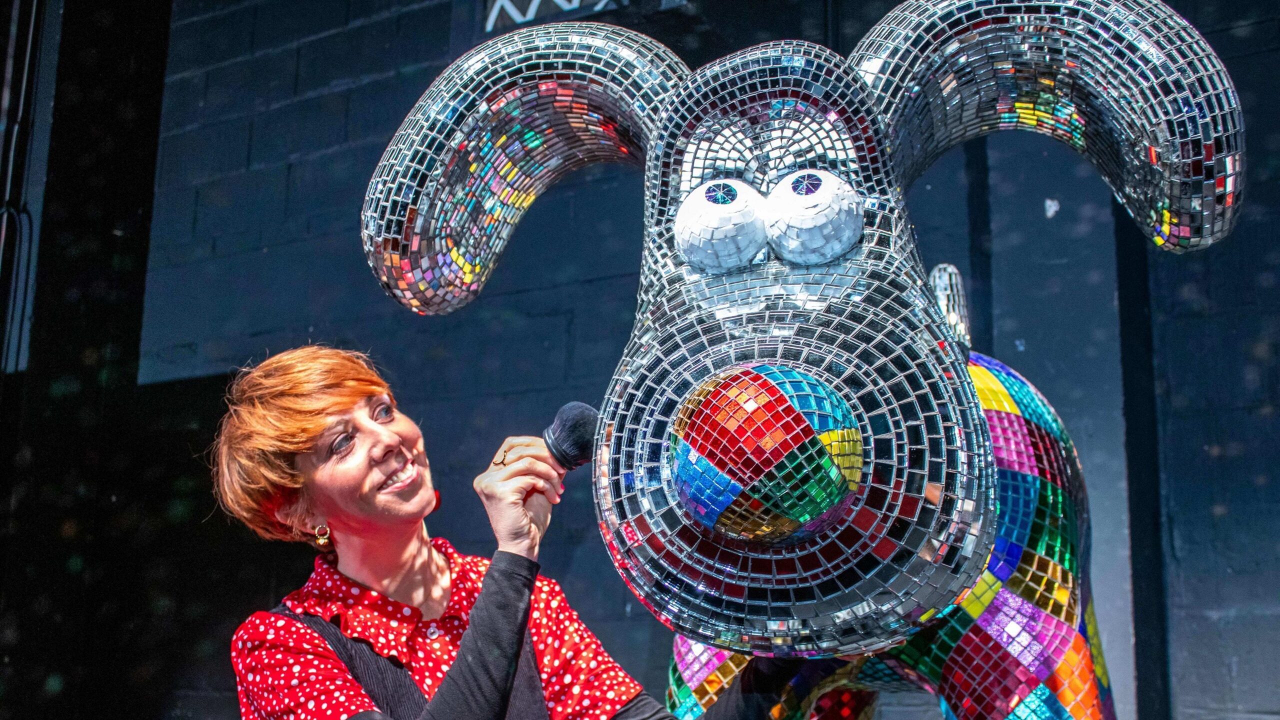 A metal multi-coloured statue of Wallace the dog. A woman is wearing a red shirt with white spots and is brushing the statue with a black brush.