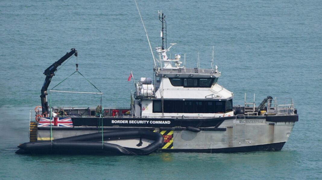 A Border Security Command vessel on the sea with a large black inflatable dinghy hanging from a crane alongside. The dinghy is several metres long and attached to the side of the boat.