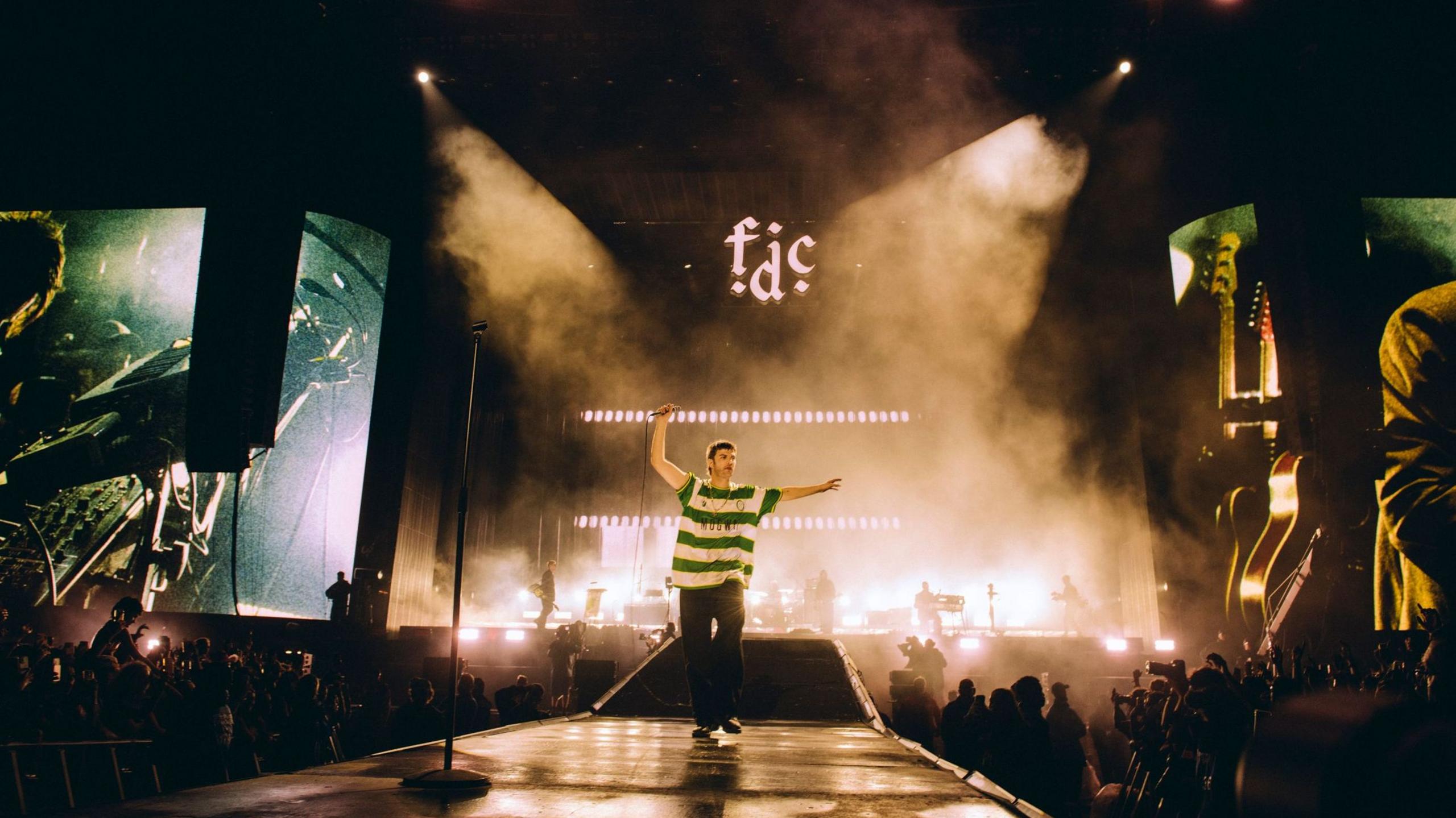 A landscape shot of Fontaines D.C. performing at Reading Festival. Grian Chatten is centre stage and seen on two large screens to each side of the stage, performing whilst wearing a Cwmbran Celtic FC away shirt.