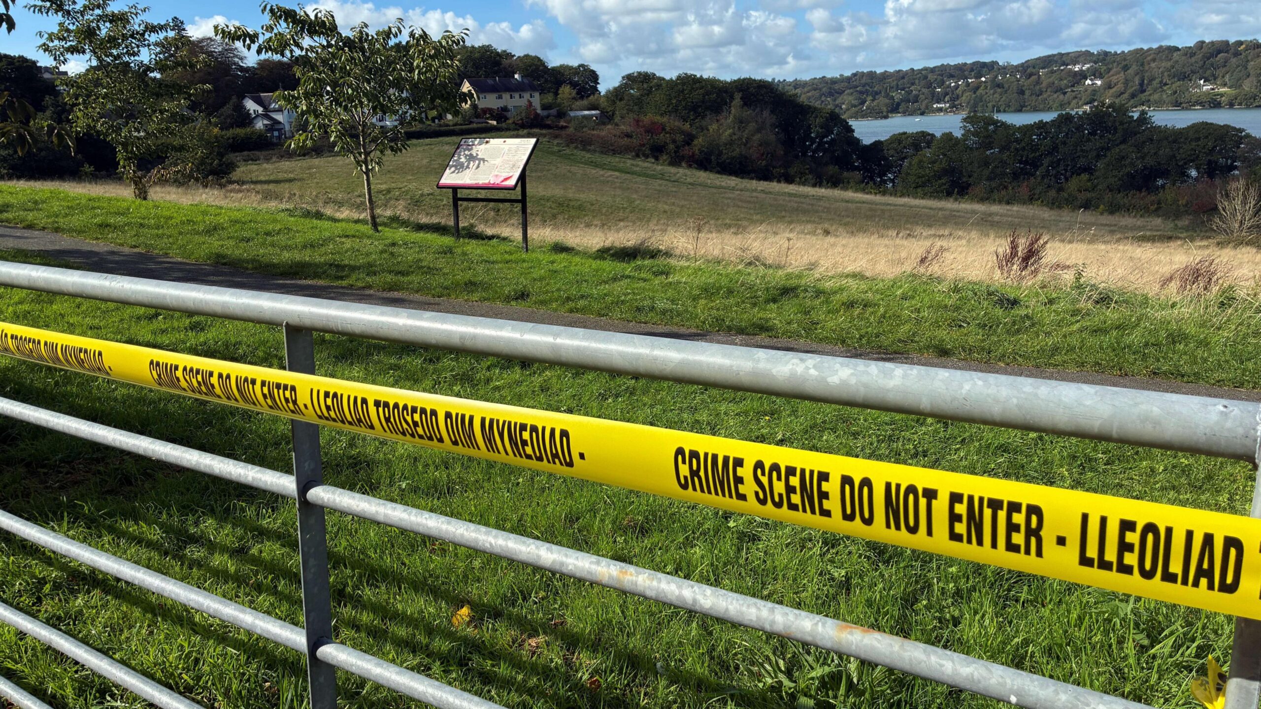 A gate has yellow crime scene tape over it. Behind the gate is a field surrounded by green trees and a river.