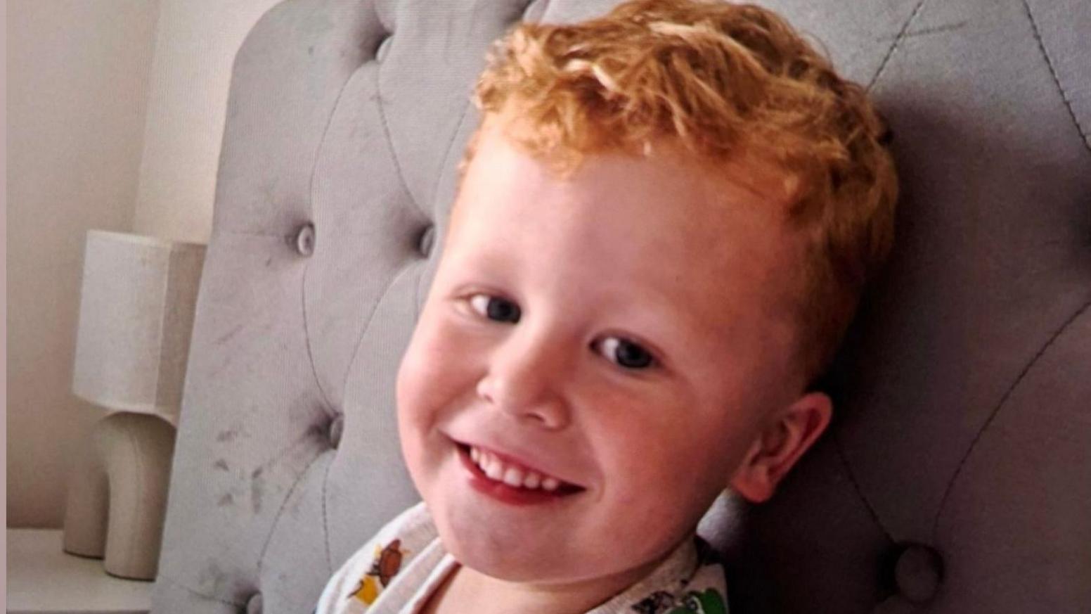 Three-year-old Noah Woods, with light hair and wearing a brightly coloured top, smiles at the camera.