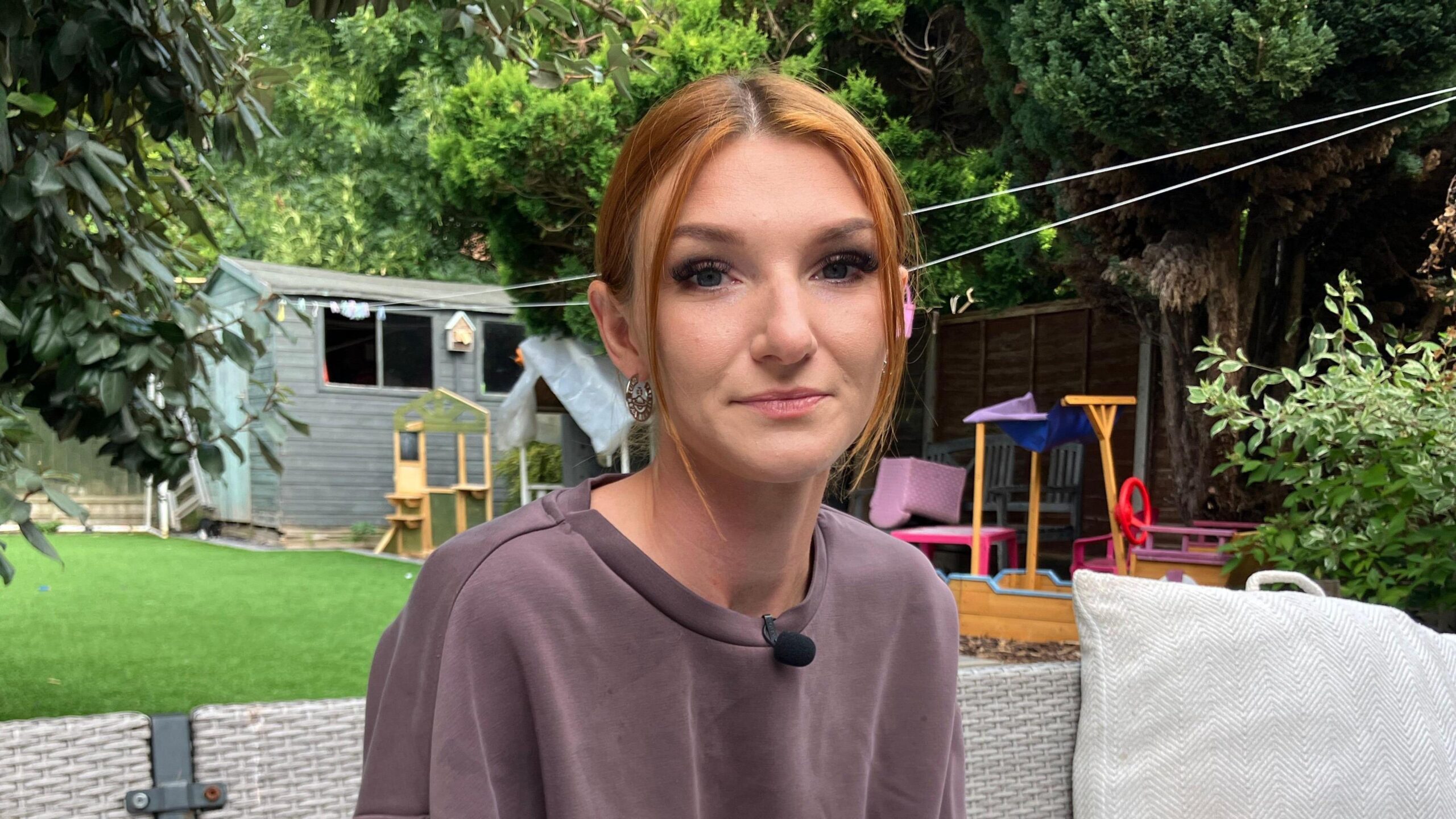 A photograph of a woman looking towards the camera. She has short hear and wears a grey crew neck jumper. She is sat outside in the garden of a property. Trees, a shed and other garden furniture can be seen in the background.