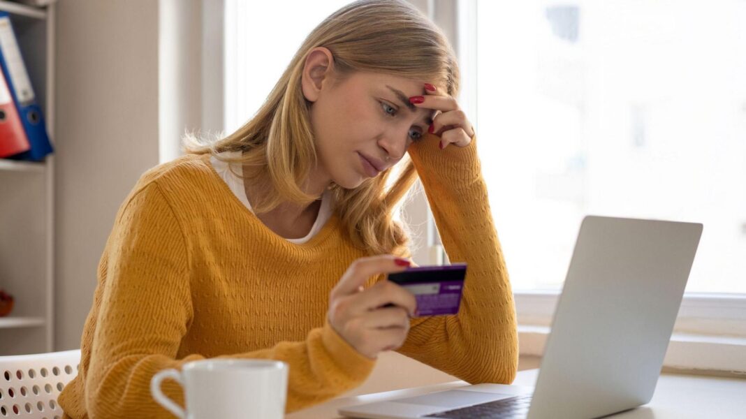 A woman sits at her laptop while holding her bank card in her right hand. She looks deep in thought, with her left hand on the side of her head. She has blonde hair and is wearing a yellow jumper.