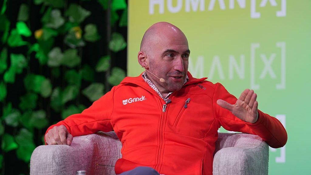 George Arison speaks onstage during the "AI, Identity, and the Future of Connection" panel at the HumanX Conference San Francisco 2026 . He wears a red casual zip up sweater with the Grindr logo in white and speaks into a microphone.