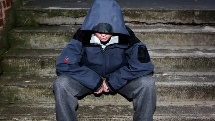 A young boy sits on some steps with a hood up, looking at the floor. He has jeans on and his face is partially hidden.