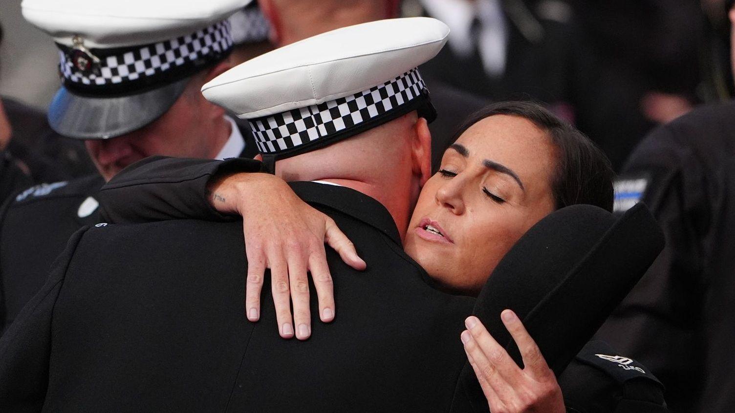 A female police officer is hugged by a male officer. She has dark brown hair which is tied back and has her eyes closed. She is holding a black hat in one of her hands. The male officer is facing away from the camera and is bald and wearing a white hat with a white and black checked trim. There are other officers around them who are out of focus.