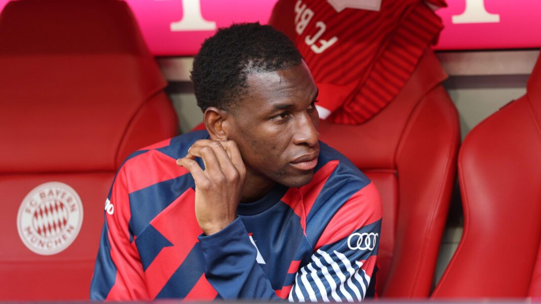 Nicolas Jackson, wearing a blue and red training top, sits on the Bayern Munich bench before a Bundesliga match against Cologne