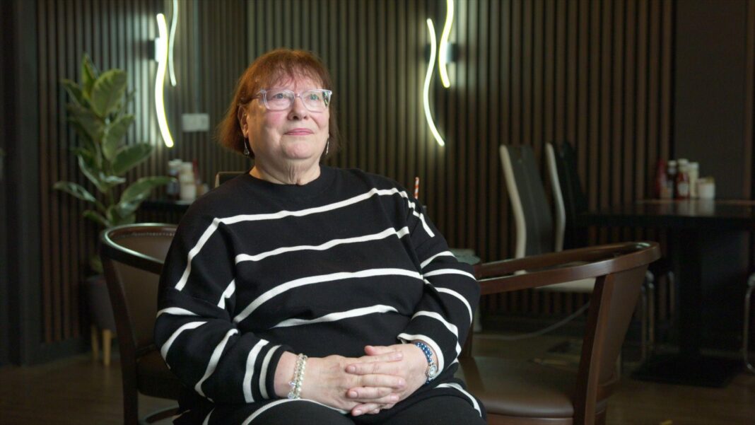 A woman with short reddish hair with a neutral expression faces the camera while sitting a room with black and brown wallpaper.