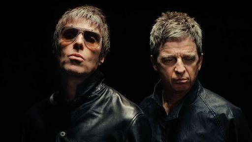 Liam and Noel Gallagher, both wearing black and looking serious in front of a black backdrop. Liam is wearing sunglasses