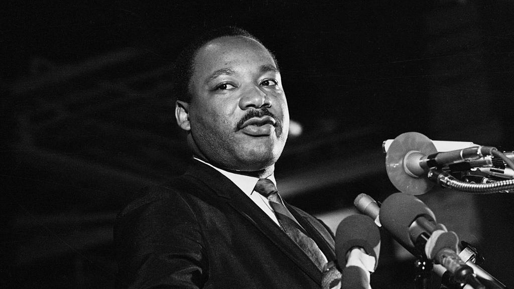 A black and white photograph of the late Dr Martin Luther King Jr. He is standing at a podium, surrounded by microphones, as he speaks, looking to the right of the stage.
