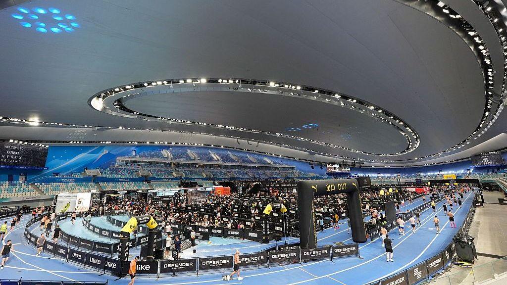 A view from the stands of Beijing's National Speed Skating Oval which is hosting a Hyrox event. There are dozens of athletes running around the track, with barriers to separate the lanes.