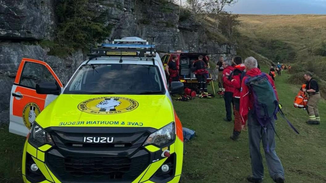 An emergency service vehicle parked on a grassy area, next to a cliff face. In the background, people wearing red waterproof jackets can be seen preparing safety equipment.