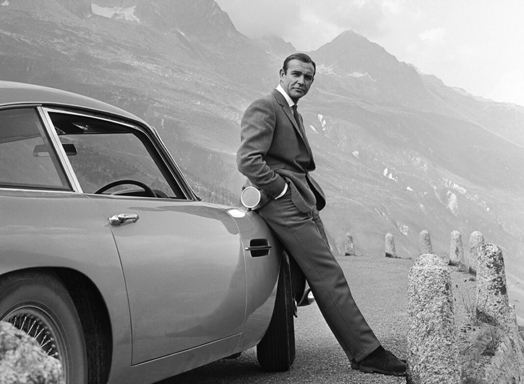 A black and white photo of a man leaning against an old car looking past the camera. He is in a suit and is parked in a mountain range.
