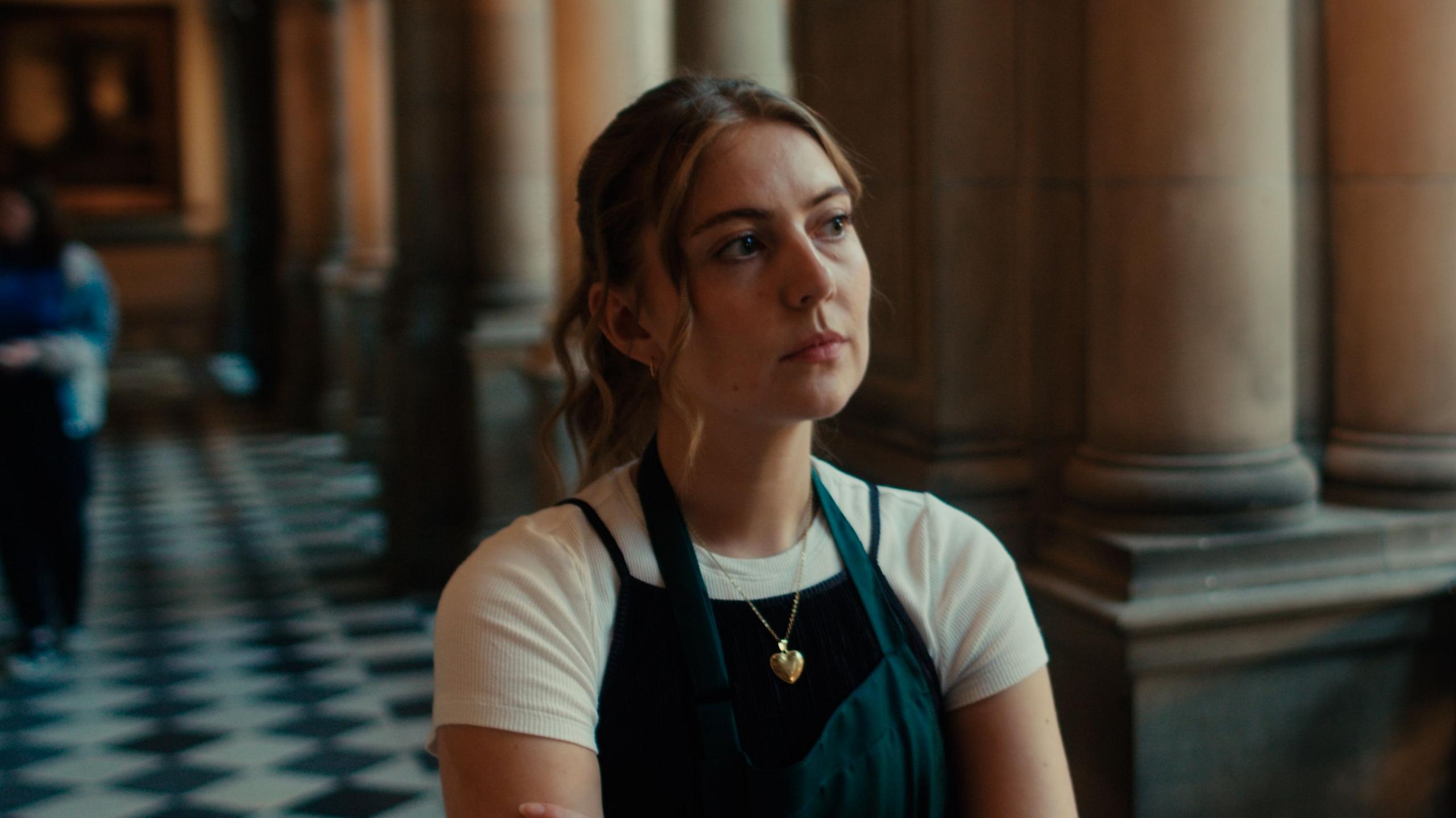 A woman wearing a dark apron and with light hair over a light-coloured shirt stands inside a grand historic building with tall stone columns and a black-and-white chequered floor. She has a serious expression from her face.