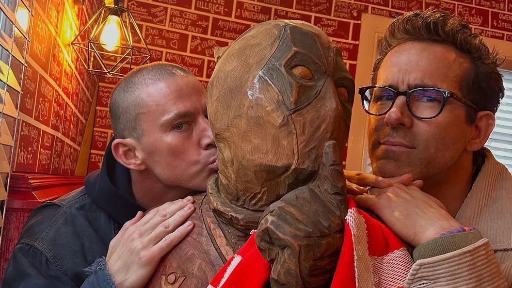 Channing Tatum and Ryan Reynolds at The Turf pub in Wrexham with a Deadpool sculpture. Tatum is kissing the cheek of the sculpture.
