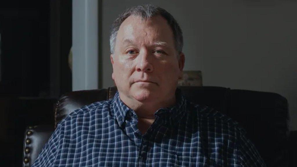 Larry Bushart sits and looks straight ahead while wearing a navy blue checkered shirt.