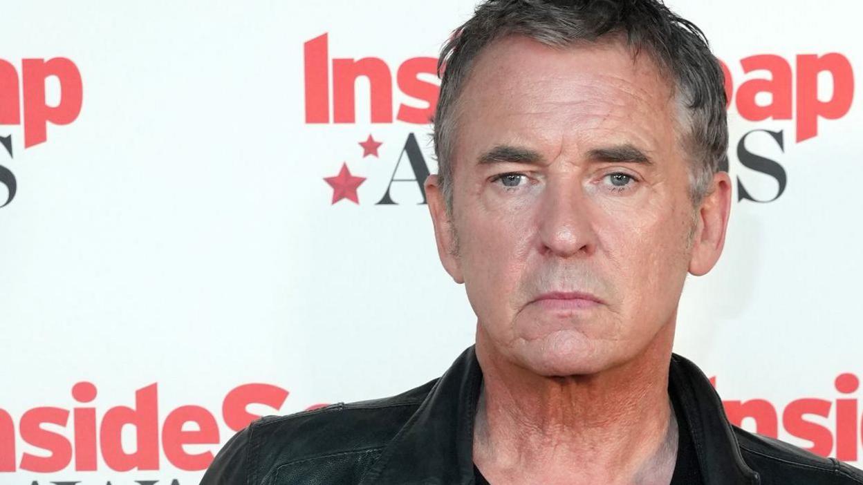 A man wearing a leather jacket and a black top standing in front of the press, there is a backdrop behind him that reads inside soap awards 2024