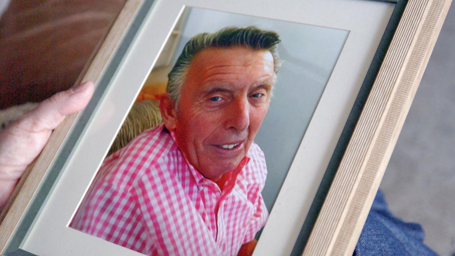 A framed colour photograph showing Graham Slinn in a pink and white checked shirt. The frame is being held by a hand visible at the edge of the image. The photograph fills most of the frame, with part of a sofa and an indoor setting visible in the background.