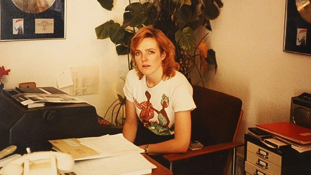 A woman sitting in an office with gold discs on the wall. She is sitting in front of a typewriter, surrounded by paper. There is a white landline phone in the foreground and a filing cabinet to the right of the picture.