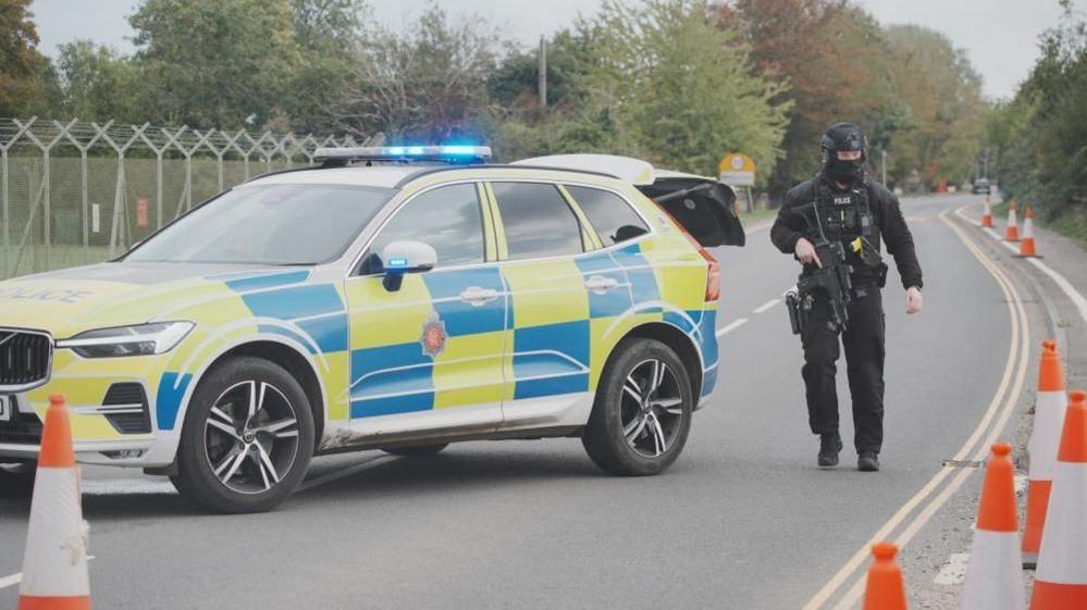 An armed specialist police officer dressed in black and a helmet and balaclava holds an automatic gun as he stands next to a police car in Fairford
