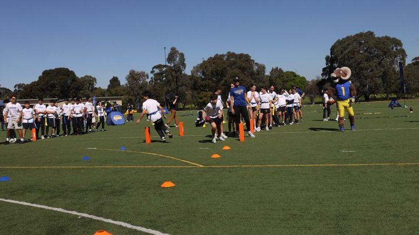 Teens dressed in white t-shirts and black shorts play ball on a sports field while a man dressed as a Rams mascot looks on