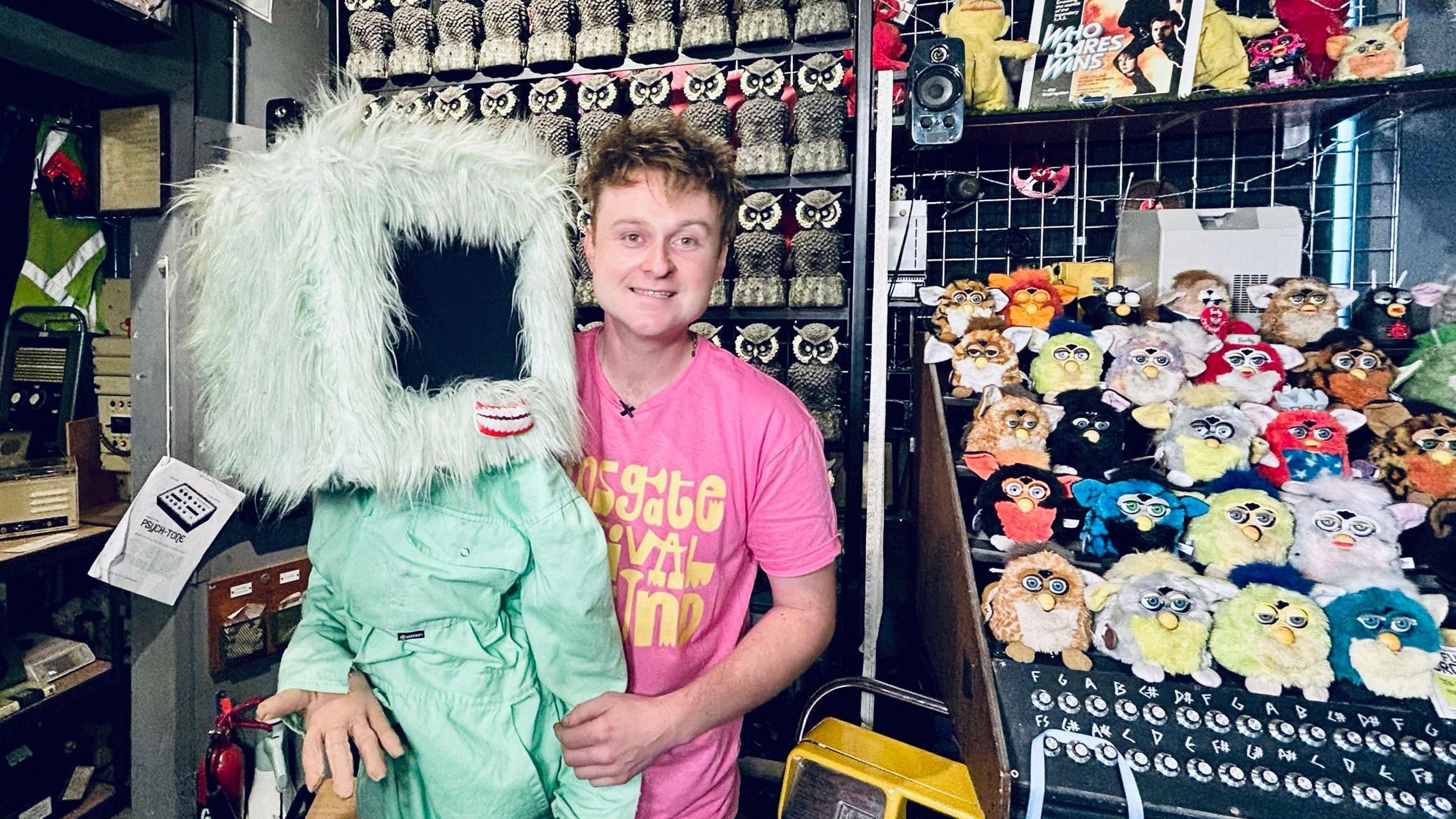 Sam Battle, wearing a bright pink T-shirt, stands beside a large pale green furry character with a rectangular black screen in place of a face. Behind them are shelves filled with colourful toys, rows of owl figures and electronic equipment. Parts of keyboards, instruments and technology displays are visible throughout the museum setting.