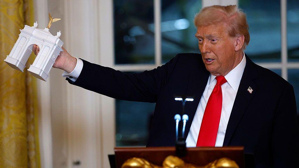 Trump holding up a tiny model of a triumphal arch in the White House
