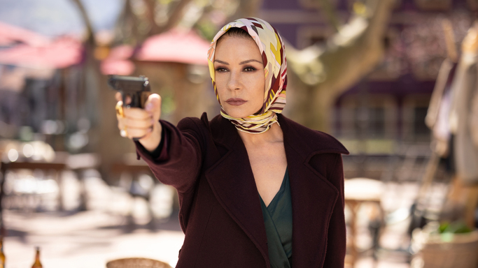 Catherine Zeta-Jones holding a gun up, wearing a scarf around her head and a maroon jacket in the Kill Jackie series.
