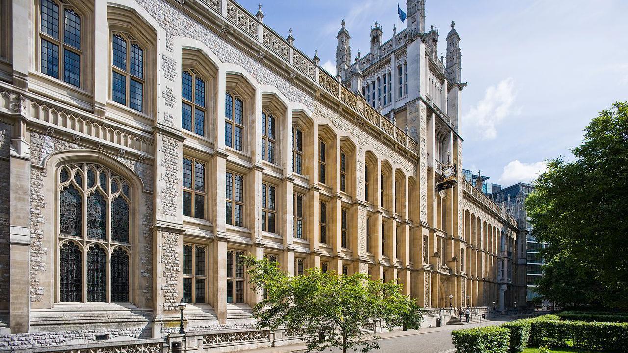 A university library which resembles a large chapel with a tower in the middle. There is a hedge and a tree on the right and a sapling in the foreground.