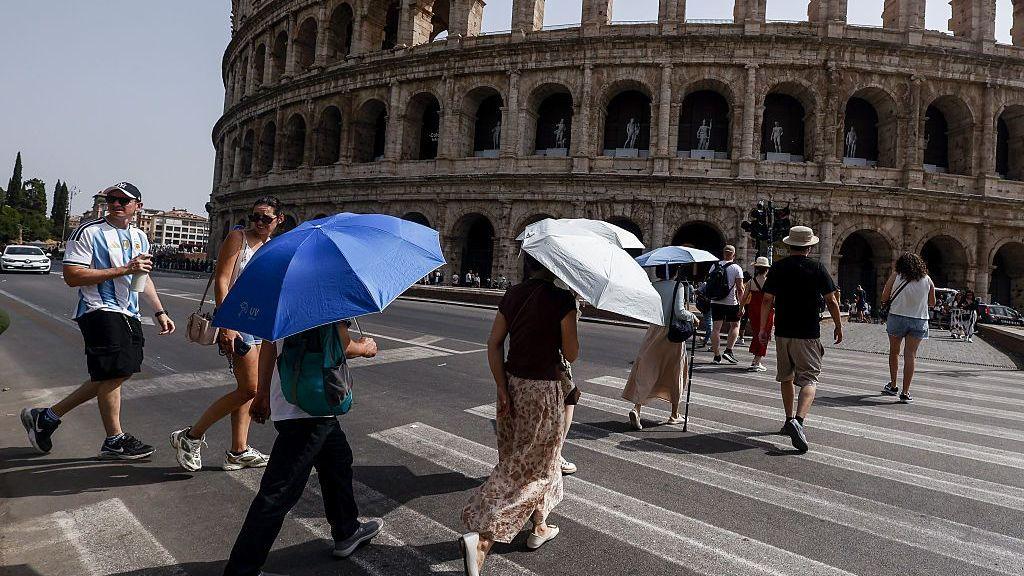 A number of people are crossing the street in Rome, in the shadow of the Colosseum,  - several are holding umbrellas and sunhats as protection from the Sun. The sky is blue and cloudless on the left of the picture some city buildings and trees can be seen.