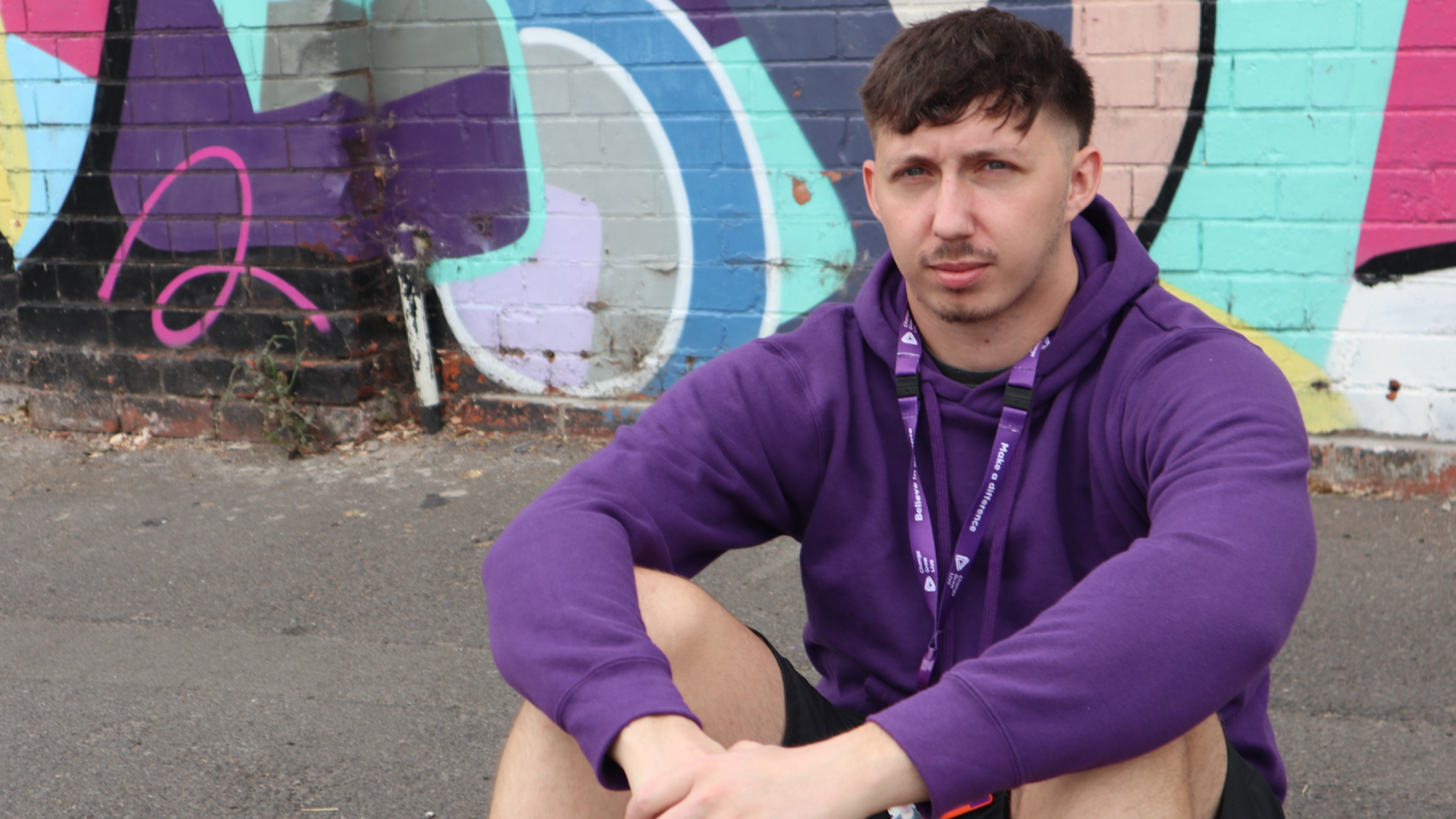 A young man with short brown hair. He is wearing a purple hooded top and is sitting on a street curb next to a graffiti-covered wall.