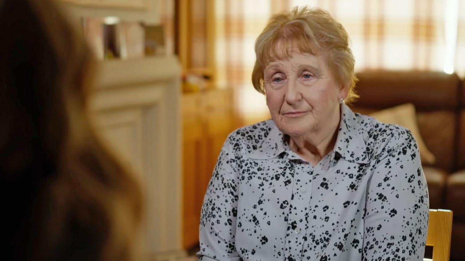 Pam Brown is seated indoors during an interview, wearing a light-coloured patterned blouse and small stud earrings. There is a person looking at her, slightly off-camera. The softly blurred background includes a sofa, curtains, and warm-toned furnishings, creating a domestic setting with natural light.