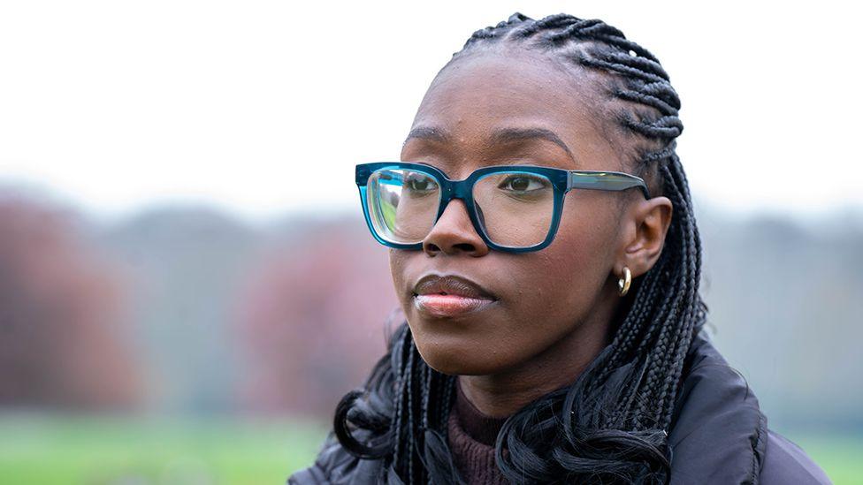 Drug gangs exploited and raped us as teens, then police failed us, women tell BBC Close-up portrait of Christine outdoors wearing large blue-framed glasses, small hoop earrings and a dark padded coat. She is looking slightly away from the camera, with braided hair visible. The background is softly blurred, showing a green field and trees in muted colours.