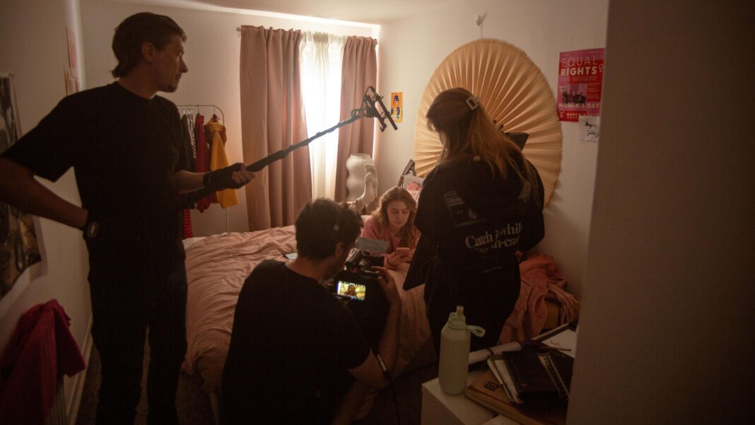 ‘Forget Notting Hill – we set our new romcom in the east end of Glasgow’ Behind-the-scenes view of a film production taking place in a small bedroom. A woman sits on a bed looking at a mobile phone while crew members surround her