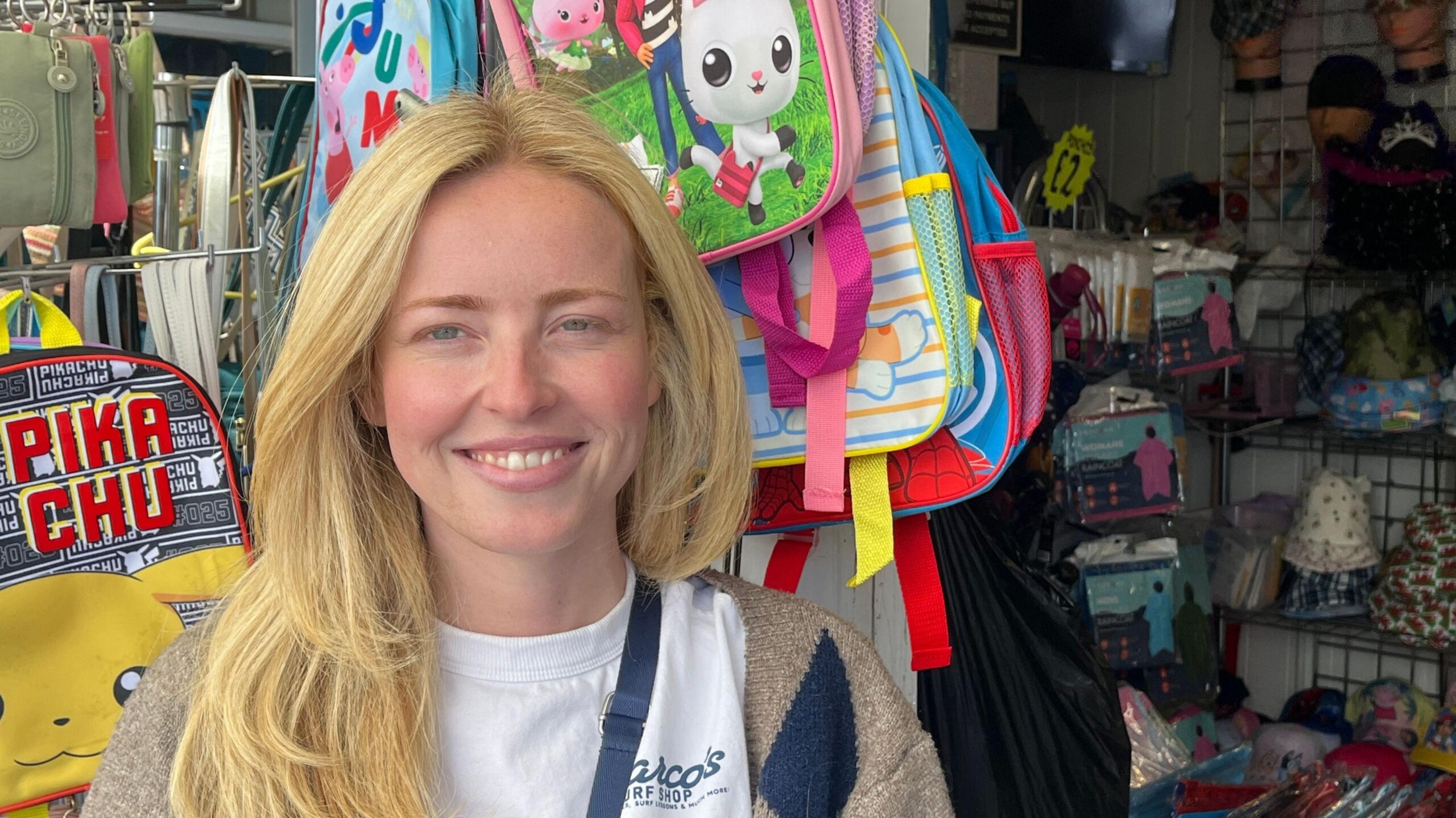 Bella, a woman with long blonde hair, smiles as she stands in front of her stall, which sells hats and bags. She is wearing a white t-shirt and a beige cardigan