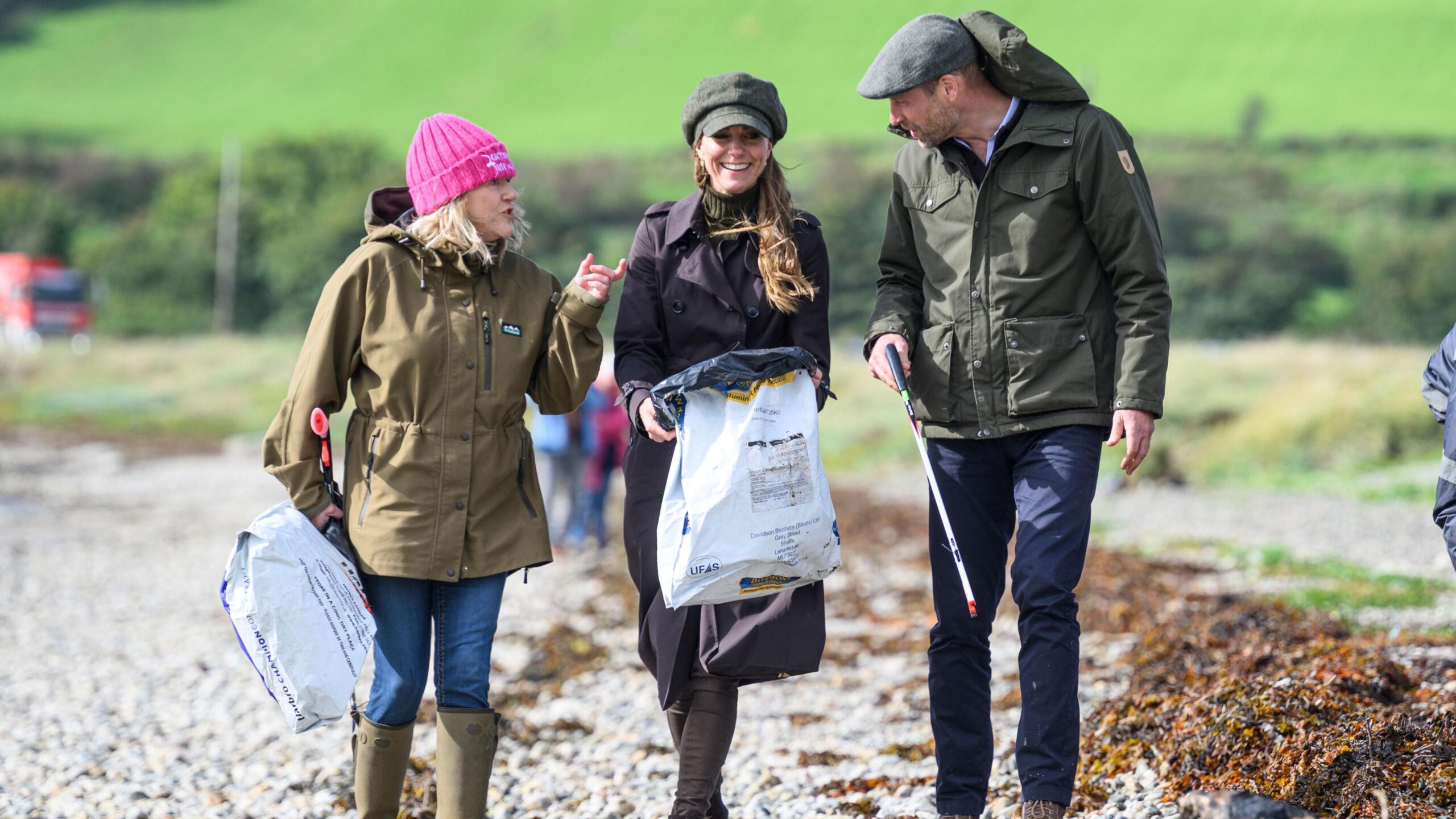 The Prince and Princess of Wales walking in coats along the beach with bags and rubbish collectors with a local wearing a pink beanie talking to them.