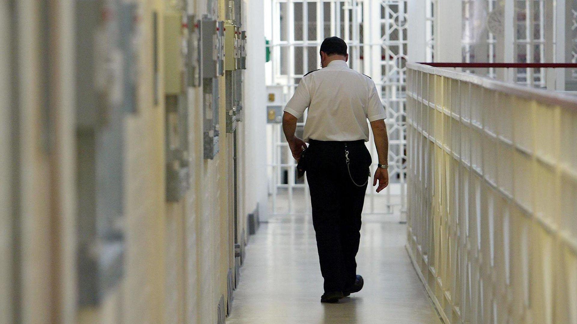 A prison officer in uniform walks past a series of cells