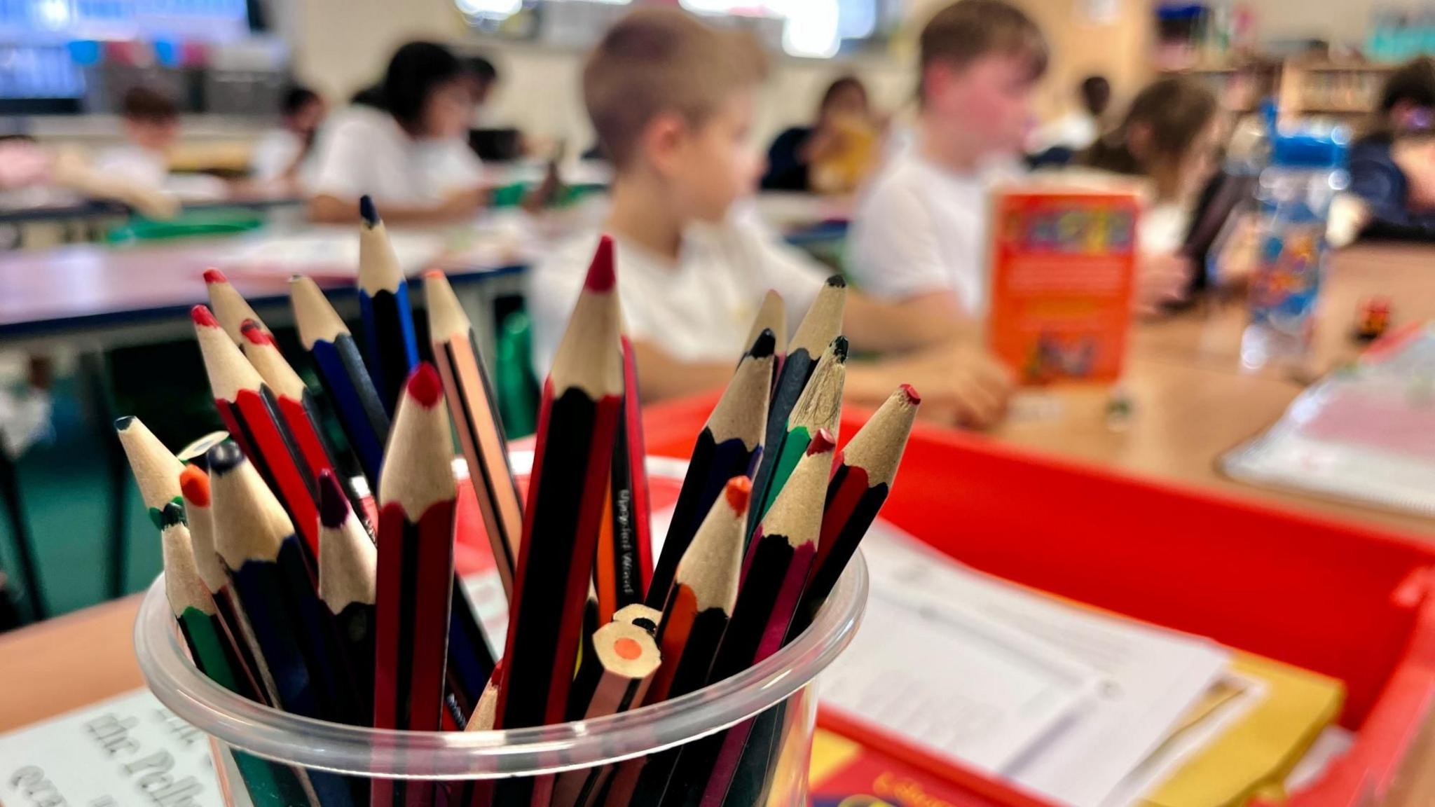 A pot of pencils is in focus in the foreground of the picture. They are on a school teachers desk with a bright red in-tray. In the background there are children sitting in rows at desks but they are out of focus and you cannot see their faces.