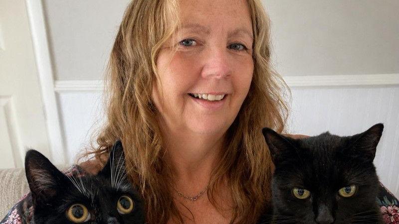 A middle-aged woman with blonde hair smiles at the camera. Two black cats are visible in front of her.
