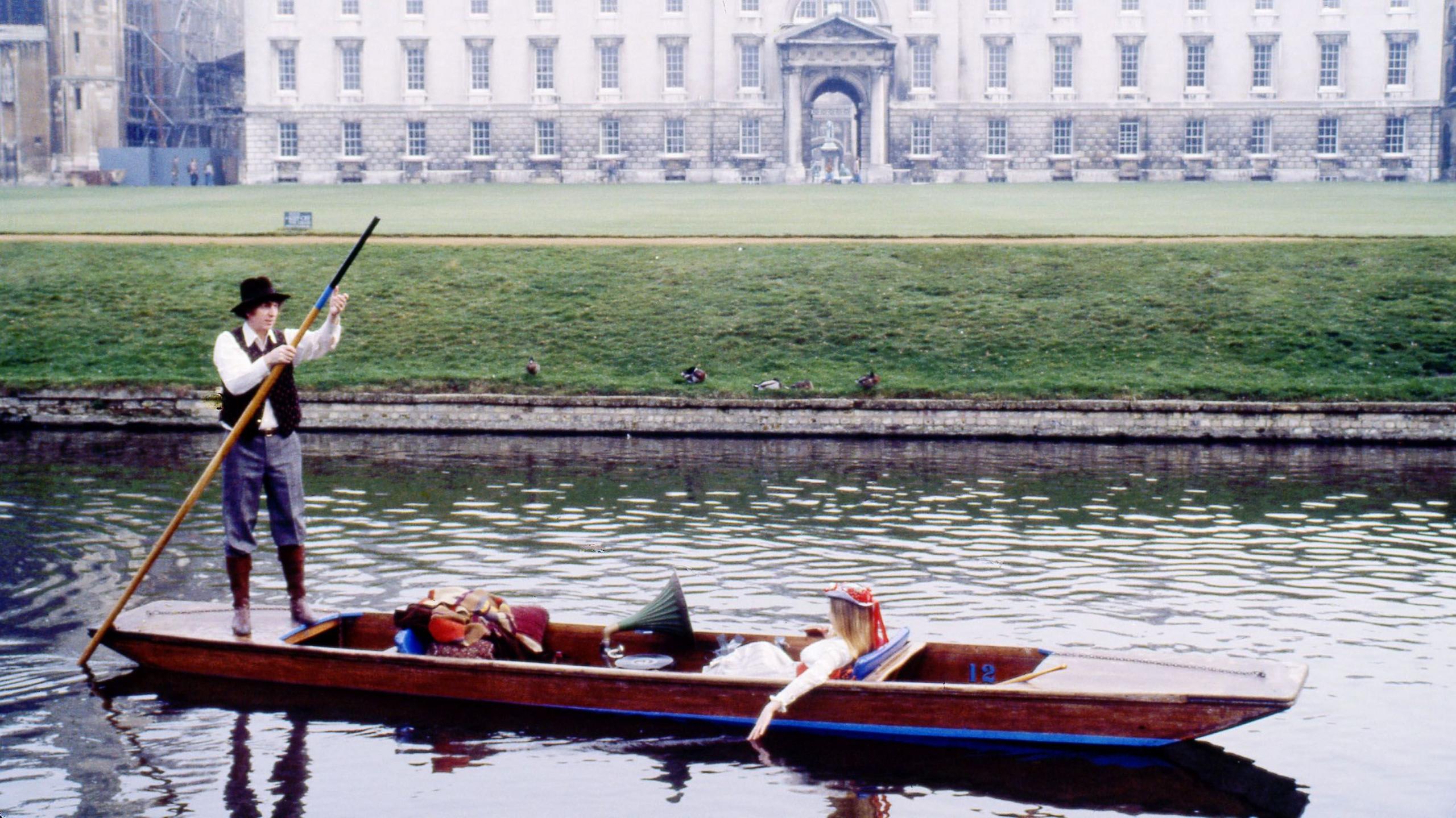 Tom Baker is wearing a black hat, white shirt and black waistcoat, navy trousers and black boots. He is standing on a wooden punt which is on the River Cam. On the right is a wall leading to a field.