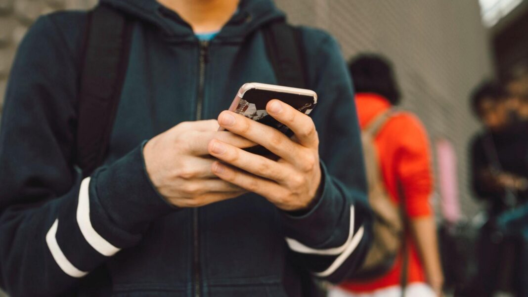 A young boy wearing a navy zip-up hoodie holds a smartphone in front of him, with other boys blurred in the background behind him.