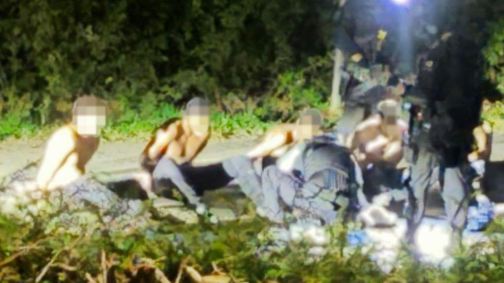 Detained men in Whelford. Their faces are blurred. They are shirtless. Trees line the background.