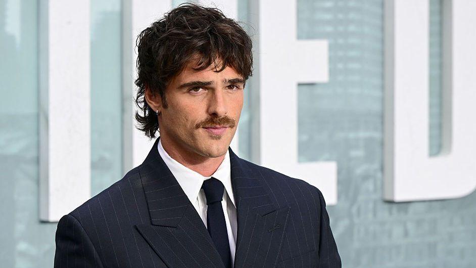 Jacob Elordi wearing a black suit, white collared shirt and black tie. He is standing on a carpet at a film premiere, looking just off to the right of the camera. He has a small silver hoop earring, thick eyebrows, a moustache and dark hair styled in a tousled mullet.
