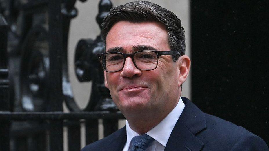 Andy Burnham, with dark speckled grey hair and black glasses. Wearing a suit, white shirt and tie.