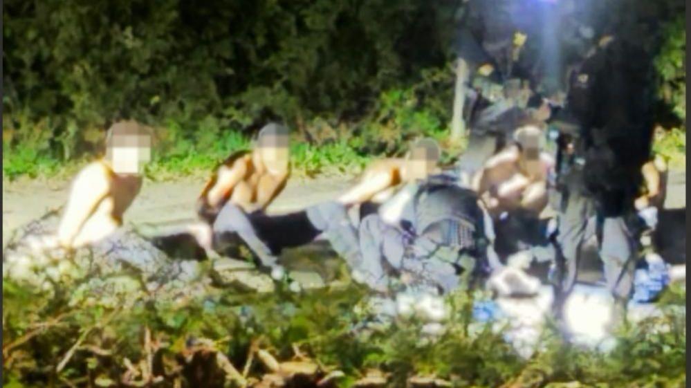 Five shirtless men sitting with their hands behind their bags as armed police surround the group. The image was taken last night and shared on a local village WhatsApp group. The original source has not been confirmed but BBC Verify has geolocated the image and can confirm it appears to be authentic