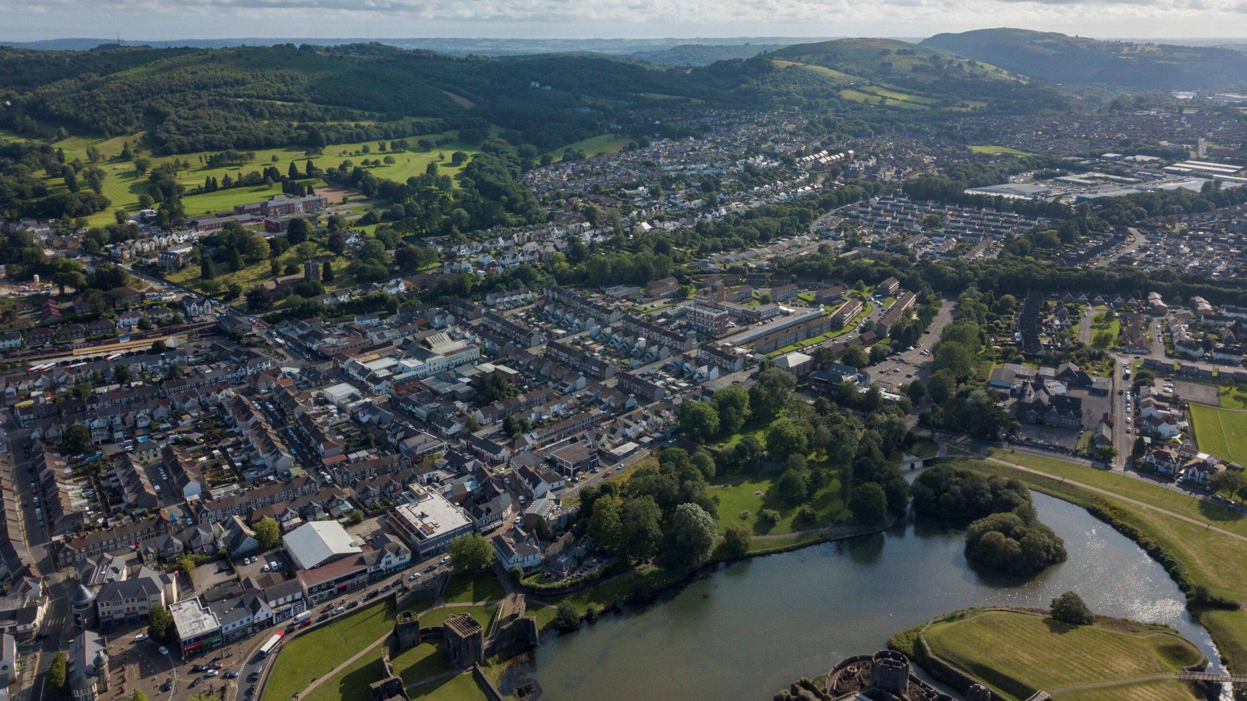 A birds eye view of Caerphilly town. It shows rows upon rows of homes surrounded by green fields and trees. There's also a big lake on the castle grounds.