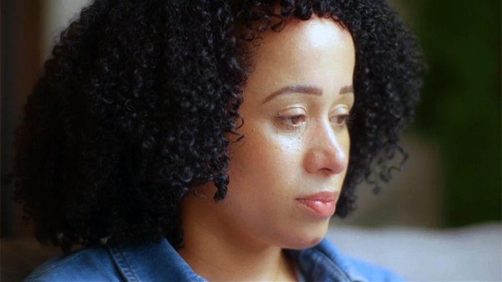 Close-up portrait of Annie, who has dark curly hair and is wearing a blue denim shirt, photographed indoors. Annie is shown in profile, looking down and away from the camera. The background is softly blurred, with warm lighting and muted green and beige tones visible behind her.